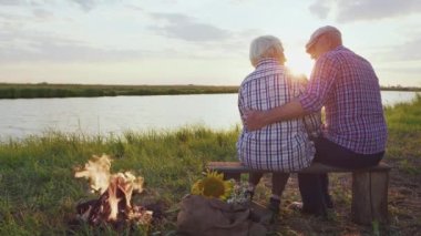 Candid feelings and embraces of two old happy lovers together sitting on lake shore near bonfire at sunset outdoors. Beauty of love, retired marriage harmony, affectionate care in carefree family life
