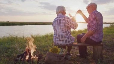 Happy old grandmother and grandfather together drink holiday beverages sitting on lake beach at sunset outdoor. Cheers anniversary celebration, friends meeting or family relax, enjoy freedom at nature
