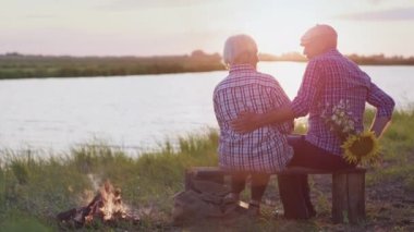 Happy affectionate husband give surprise flowers bouquet lovely amazed wife sitting on lake beach at evening walk outdoors. Joyful life moments, family holiday celebration together at summer nature