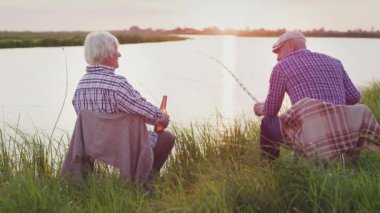 Evening family relax : happy grandparent couple enjoy vacation sitting on lake shore at sunset outdoors. Spending time fishing together, calm carefree summer leisure at nature. Love, peace and freedom
