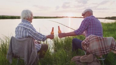 Clink bottles - cheers holiday. Two happy old people celebration weekend sitting on lake beach, enjoy sunset scene outdoors. Leisure activity together at nature, family spending time on summer evening