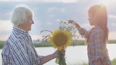 Candid feelings and embraces of two old happy lovers together sitting on lake shore near bonfire at sunset outdoors. Beauty of love, retired marriage harmony, affectionate care in carefree family life