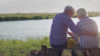 Back view at happy bonding grandfather, grandmother together hugging sitting on lake shore outdoors close up. Carefree care cuddle, candid affectionate emotions, joyful life love of old smiling couple