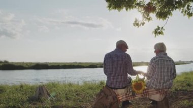 Old grandfather and grandmother together talking sitting on pond shore in sunshine outdoors. Happy loving relationship of retired couple, peaceful carefree meeting friends, calm relax walk at nature