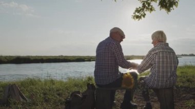 Old affectionate gentle husband kiss hand lovely wife sitting on bench on lake beach outdoor. Happy married retired couple rest together at nature, calm tenderness and care, elderly human relationship