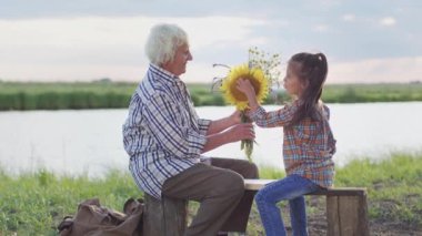 Happy family relax: old affectionate grandmother help curious small granddaughter flower beauty sitting on lake beach outdoors. Enjoy peaceful environment together, teach children art and nature love