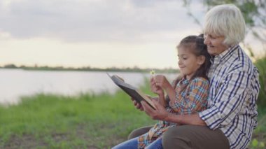 Happy smiling old grandmother reding funny book fairy tale, embracing little joyful granddaughter sitting on lake beach outdoors. Child thinking and imagination development, calm family rest at nature