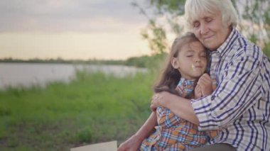 Happy smiling child granddaughter and old affectionate grandmother together embracing sitting on lake shore outdoors. Candid human feelings, family love and care, carefree childhood in harmony, peace