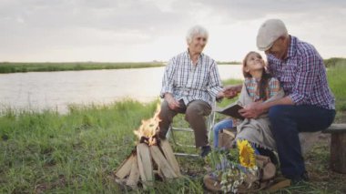 Happy smiling family spend time together outdoor : old caring grandmother read book story granddaughter at sunset. Relationship harmony of grandparents and children, imagination fantasy, peaceful life