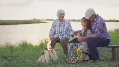 Happy smiling family spend time together outdoor : old caring grandmother read book story granddaughter at sunset. Relationship harmony of grandparents and children, imagination fantasy, peaceful life