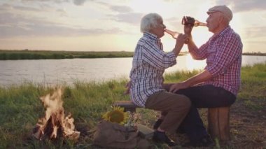 Happy old couple of grandfather and grandmother embraces sitting on lake shore outdoors. Affectionate tender feelings, romantic relationship of elderly spouses spending time together at summer nature