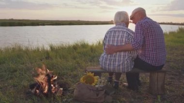 Candid feelings and embraces of two old happy lovers together sitting on lake shore near bonfire at sunset outdoors. Beauty of love, retired marriage harmony, affectionate care in carefree family life