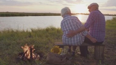 Rear view at two old hugging spouses sitting at lake beach enjoying love at sunset outdoors. Happy family rest walk together, older marriage harmony, gentle human relationships, joyful life together
