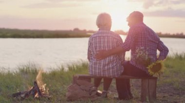 Happy old affectionate couple holding hands sitting on summer date enjoying rest on lake shore outdoors. Real life love, human care feelings and romantic relationship, spending time together at nature