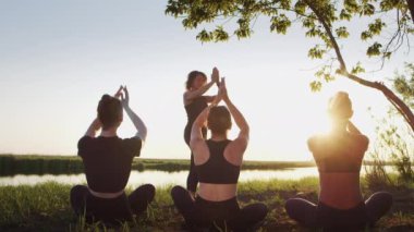 Üç dişiden oluşan bir grup siluet, dışarıda, alacakaranlıkta sakin suyun yanında nilüfer çiçeği pozisyonunda oturarak yoga yapıyor. Duygusal rahatlama için sakin namaste, stres yok.