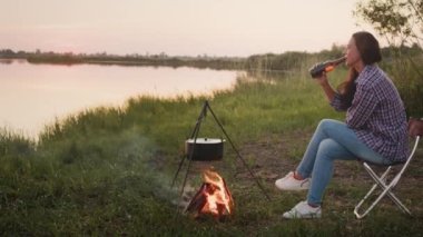 Clink bottles - cheers holiday. Happy young person celebration weekend sitting on lake beach, enjoy sunset scene outdoors. Leisure activity together at nature, family spending time on summer evening