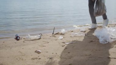 Female volunteer picks up trash left on wet sand on beach near sea on sunny day. Young woman in rubber gloves cleans area of coastal town trying to avoid environmental pollution and social problems