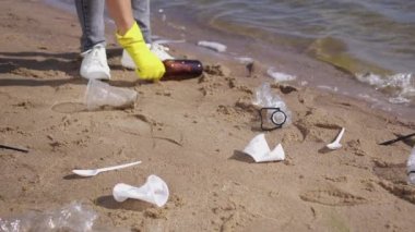 Female volunteer picks up scattered rubbish thrown on river beach on sunny day. Woman in rubber gloves walks on wet sand collecting trash into blue bag to reduce environmental pollution closeup