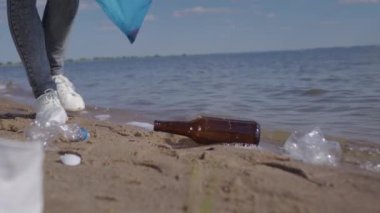 Woman picks up plastic bags and bottles scattered on wet sand of seashore to avoid water pollution. Female activist takes care of environment trying to protect beach from contamination closeup