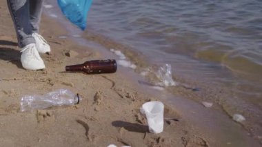 Woman picks up plastic bags and bottles scattered on wet sand of seashore to avoid water pollution. Female activist takes care of environment trying to protect beach from contamination closeup