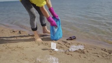 Woman in rubber gloves walks on beach with wet sand picking up scattered plastic rubbish to avoid water pollution. Female person cleans seashore on sunny day taking care of environment and nature