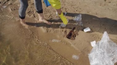 Female volunteer tries to avoid water contamination picking up trash on wet sand and putting into plastic bag. Barefoot woman cleans public beach taking care of environment on sunny day closeup