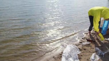 Woman takes care of environment and ecology picking up plastic rubbish scattered on wet sand of public river beach on sunny day. barefoot lady volunteer cleans areas trying to avoid water pollution