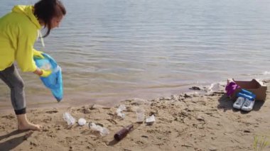 Woman takes care of environment and ecology picking up plastic rubbish scattered on wet sand of public river beach on sunny day. barefoot lady volunteer cleans areas trying to avoid water pollution