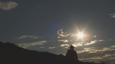 Man in hat goes uphill at back sunset light in countryside enjoying hiking in evening. Dark silhouette of male hiker leading active lifestyle exploring wild nature against bright sun on cloudy sky
