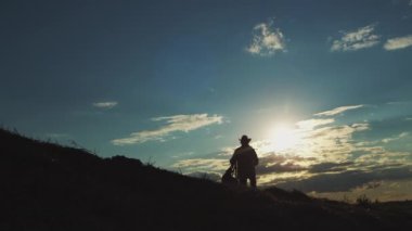 Sportive man walks in highland at bright back sunset light exploring wild nature in evening. Dark silhouette of male person in hat walking in rural area against illuminated cloudy sky backside view