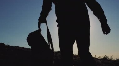 Dark silhouette of man walking in highland putting on backpack at back sunset light. Adventurous male person explores wild nature while hiking in rural area against clear blue sky backside view