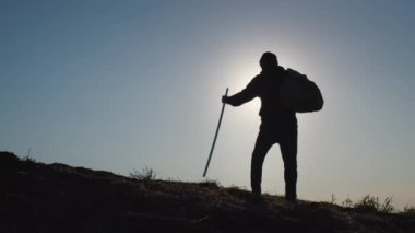 Adventurous man stands on high hill spreading hands to sides with joy at back light after throwing trekking pole and backpack on ground. Silhouette of traveler against blue sky at sunset backside view