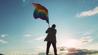 Proud LGBT representative lifts varicolored flag at sunset standing on top of hill against illuminated cloudy sky. Dark silhouette of man with banner at bright back light in wild area backside view