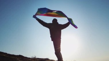 Proud man in cap stands on top covered with varicolored flag at back light after climbing up hill. Male person lifts flag to show support for LGBT community against cloudy sky at sunset backside view