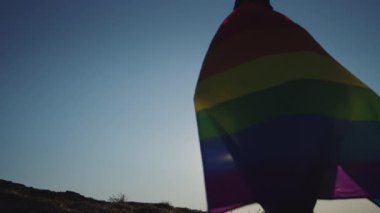 Young man goes uphill in wild area covered with rainbow LGBT flag in evening at bright back light. Proud LGBT representative silhouette raises banner against clear blue sky at sunset backside view