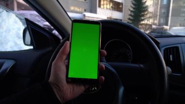 Arabadaki sürücü şehir bahçesinde yeşil ekranlı bir telefon tutuyor.