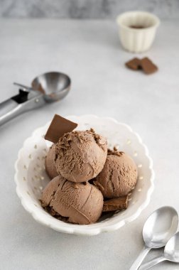 Üç kahverengi çikolatalı dondurma topu, gri arka planda beyaz kasede kepçe.