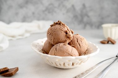 Üç kahverengi çikolatalı dondurma topu, gri arka planda beyaz kasede kepçe.