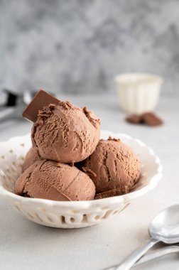 Üç kahverengi çikolatalı dondurma topu, gri arka planda beyaz kasede kepçe.