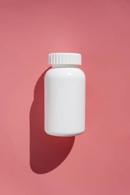 Beyaz plastik hap şişesi, vitamin ya da gıda takviyesi. Pembe arka planda ilaç hapları olan, güneş ışığı ve gölgeli bir kap.