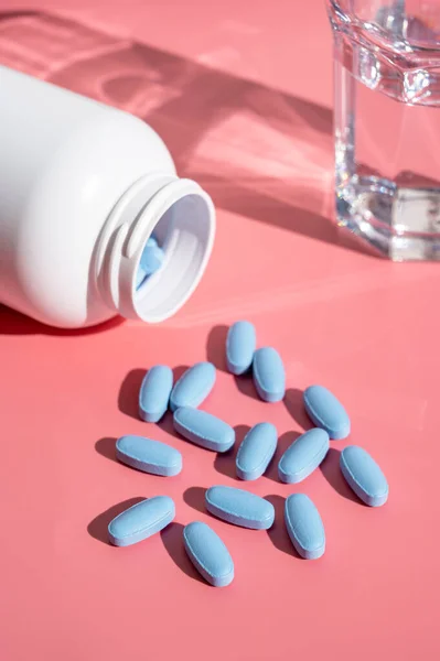 Natural food supplements, blue pills and glass of water on pink background, sun light with shadows