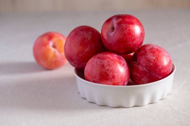Masanın üzerinde beyaz bir seramik kasede açık keten masa örtüsü olan kırmızı erik. Sağlıklı organik hasat