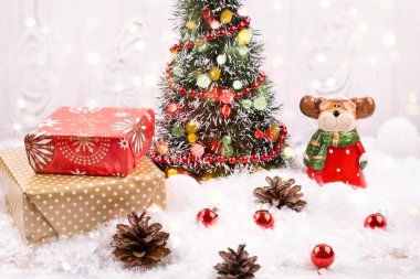 Noel arkaplanda Noel Baba gibi giyinmiş bir geyik, boncuklarla süslenmiş Noel ağacı, kağıda sarılı hediyeler, karda çam kozalağı, şenlikli arka plan, yakın plan, seçici odak.