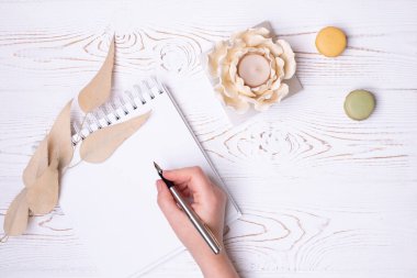 A female hand holds a pen over a clean diary with spring, macaroons, beige leaves and a flower-shaped candlestick on a white shabby wooden table. Fashion flat lay.