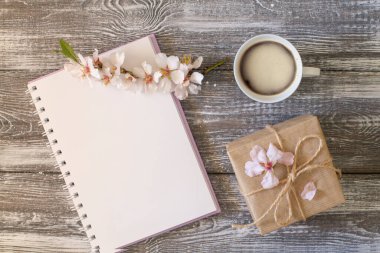 Boş günlükle bahar kompozisyonu, bir fincan kahve, kraft kağıdına sarılı hediye, bahar çiçekli badem dalı gri ahşap bir masada çiçekler. Düz yat, boşluğu kopyala