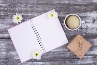 Fikirler, planlar, bir fincan kahve, kraft kağıdına sarılmış hediye ve gri ahşap masada beyaz kasımpatı çiçekleri için boş günlüğün en üst görüntüsü. Boşluğu kopyala
