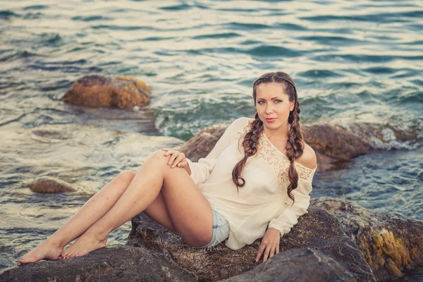 Beautiful young woman sitting on the stone on the seashore — Stock Photo, Image