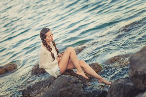 Beautiful young woman sitting on the stone on the seashore — Stock Photo, Image