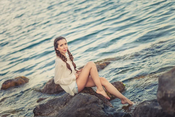 Beautiful young woman sitting on the stone on the seashore — Stock Photo, Image