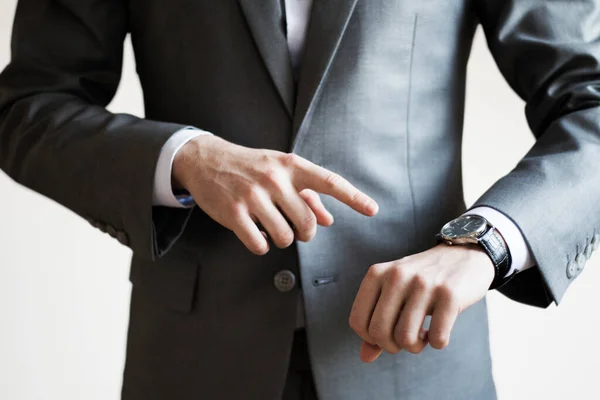 Close up of a man in grey suit pointing at watch on his hand with white background